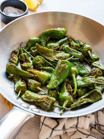 Blistered Padrón peppers with sea salt in a stainless steel pan, with lemon wedges and a bowl of coarse salt.