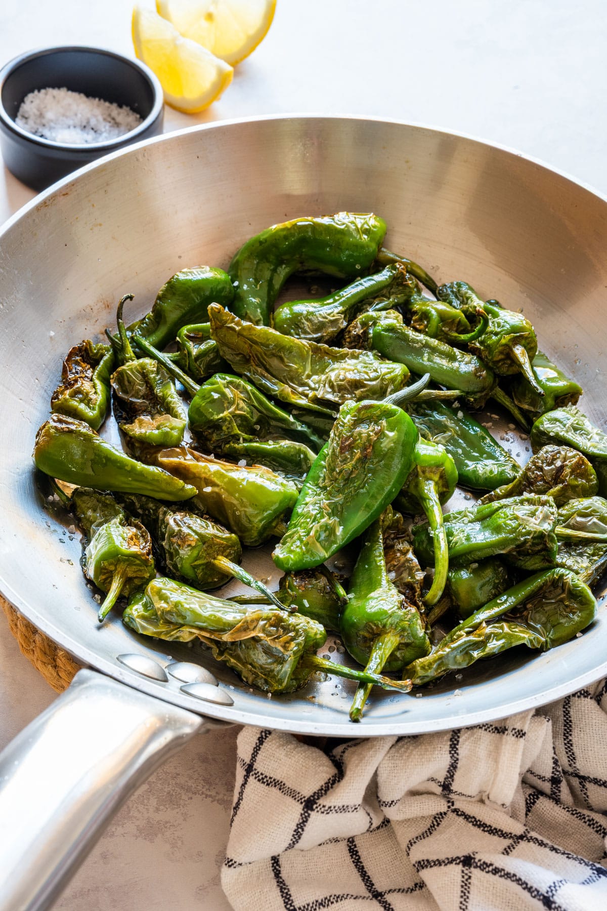 Blistered Padrón peppers with sea salt in a stainless steel pan, with lemon wedges and a bowl of coarse salt.
