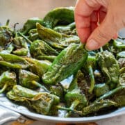 Hand holding a cooked Padrón pepper over a pan of peppers sprinkled with coarse sea salt.