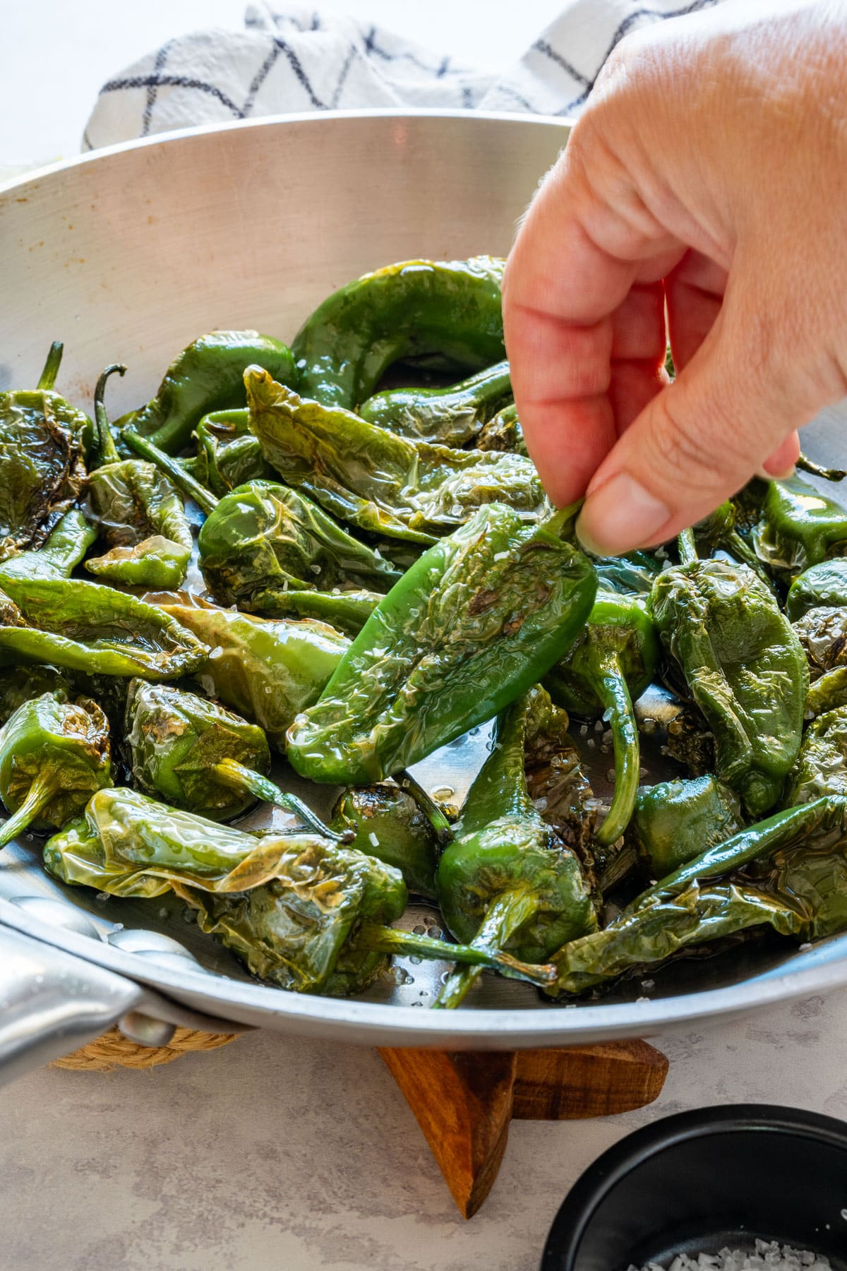 Hand holding a cooked Padrón pepper over a pan of peppers sprinkled with coarse sea salt.