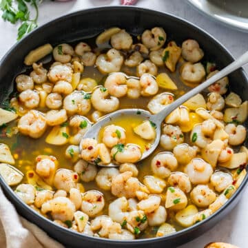 Small shrimp cooked in garlic and olive oil, lifted on a spoon over a black pan.