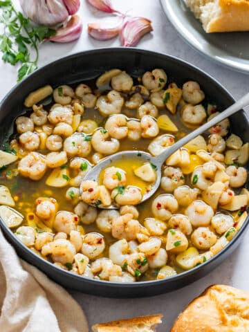 Small shrimp cooked in garlic and olive oil, lifted on a spoon over a black pan.