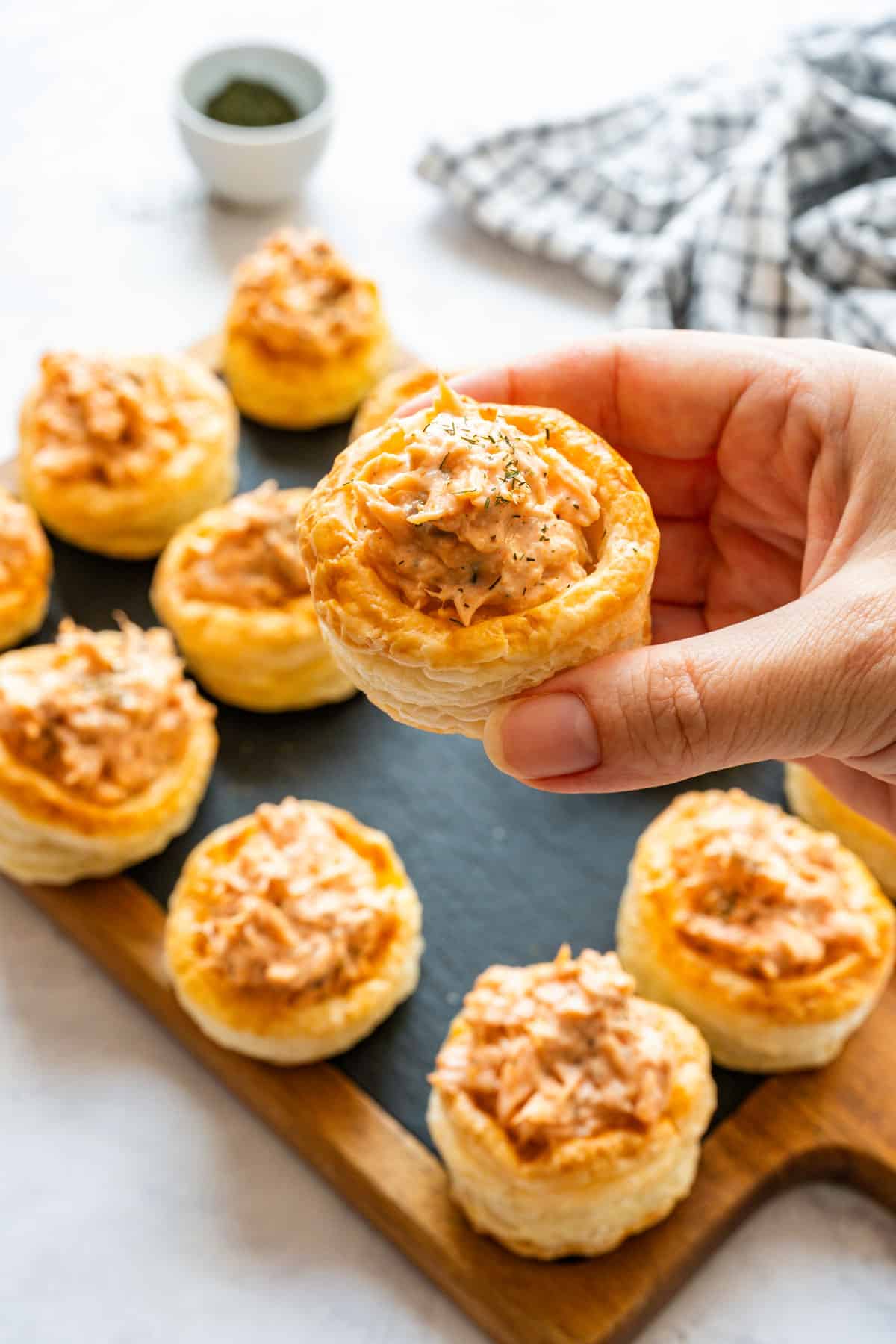 Hand holding a filled puff pastry case above a board of finished salmon appetizers.