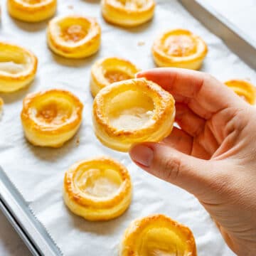 Hand holding a baked vol-au-vent case with a hollow center above a tray of more cases on parchment paper.