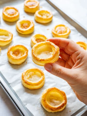 Hand holding a baked vol-au-vent case with a hollow center above a tray of more cases on parchment paper.