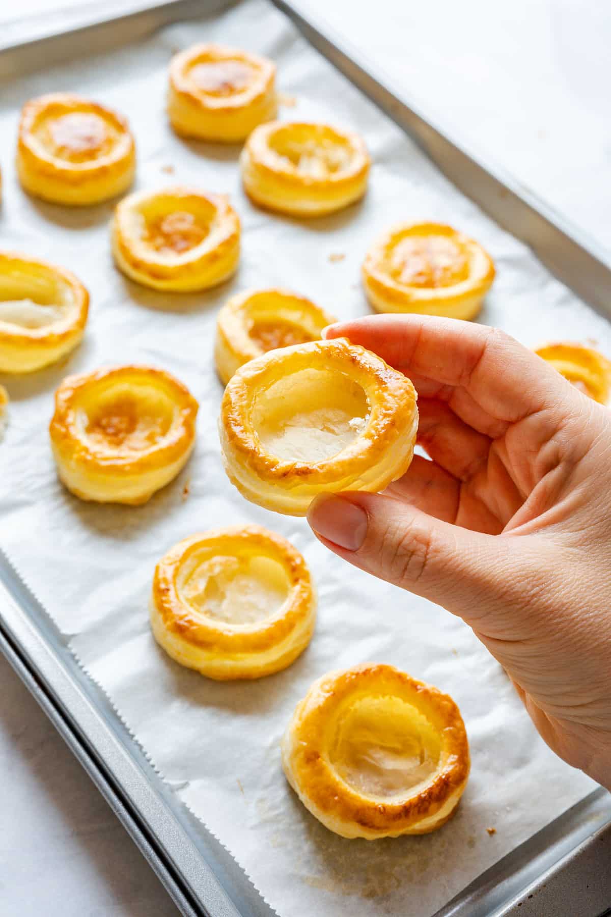 Hand holding a baked vol-au-vent case with a hollow center above a tray of more cases on parchment paper.