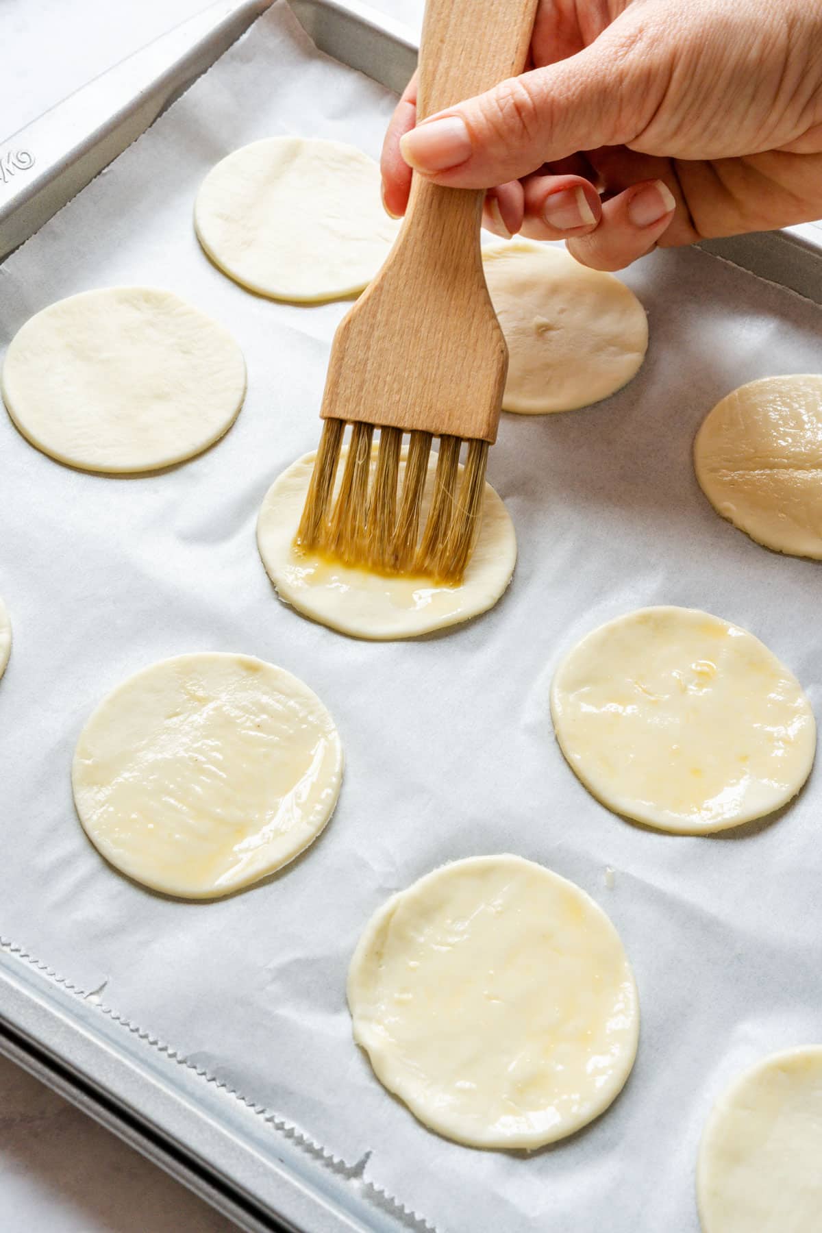 Brushing beaten egg over puff pastry circles on a parchment-lined baking sheet.