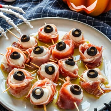 Halloween mozzarella eyeballs wrapped in prosciutto on a plate with olive oil, with a lit jack-o'-lantern behind them.