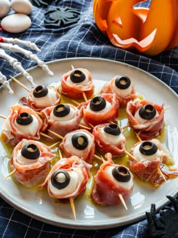 Halloween mozzarella eyeballs wrapped in prosciutto on a plate with olive oil, with a lit jack-o'-lantern behind them.