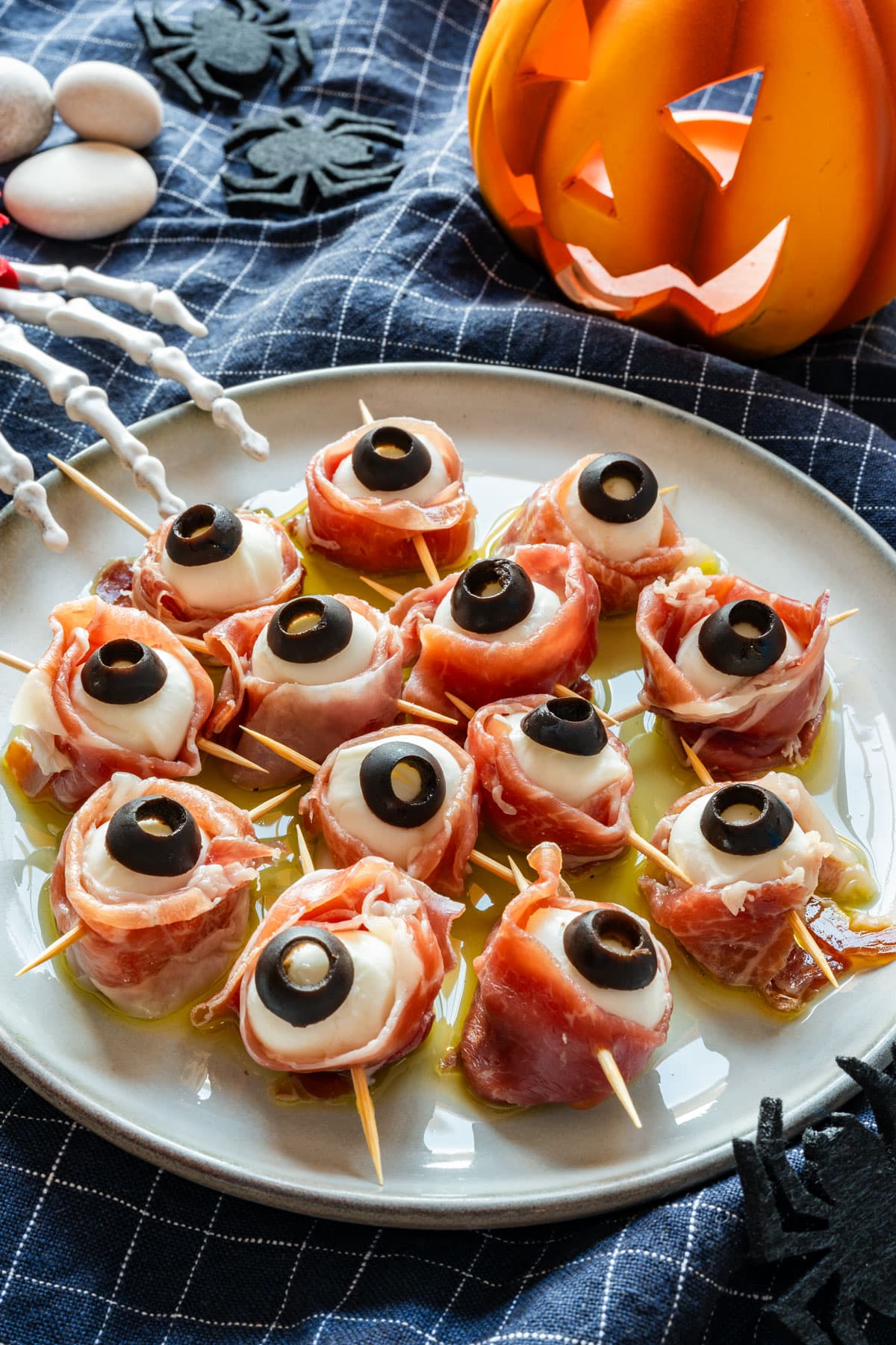 Halloween mozzarella eyeballs wrapped in prosciutto on a plate with olive oil, with a lit jack-o’-lantern behind them.