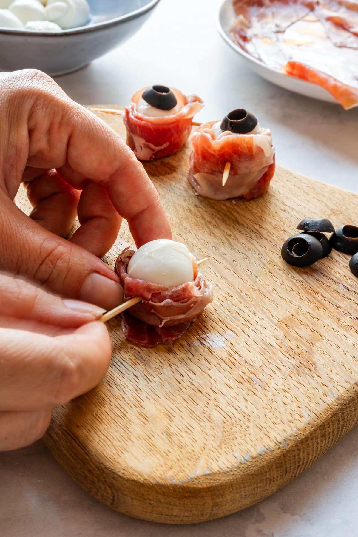 Pushing a toothpick through the prosciutto near the base of a mozzarella ball, with two finished eyeballs behind it.