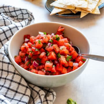 White bowl of watermelon salsa with tortilla chips behind it and fresh cilantro on the counter.
