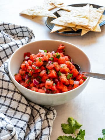 White bowl of watermelon salsa with tortilla chips behind it and fresh cilantro on the counter.