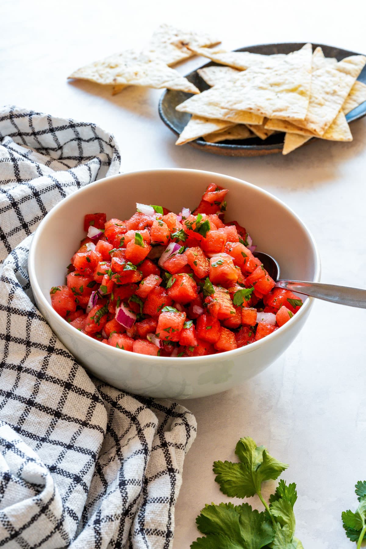 White bowl of watermelon salsa with tortilla chips behind it and fresh cilantro on the counter.