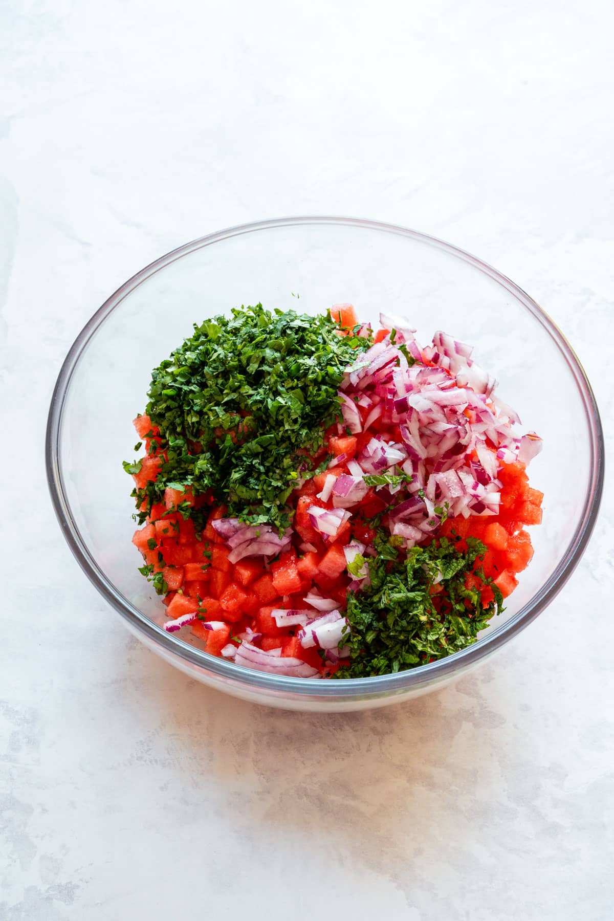 Diced watermelon, chopped red onion, cilantro and mint in a glass bowl before stirring.