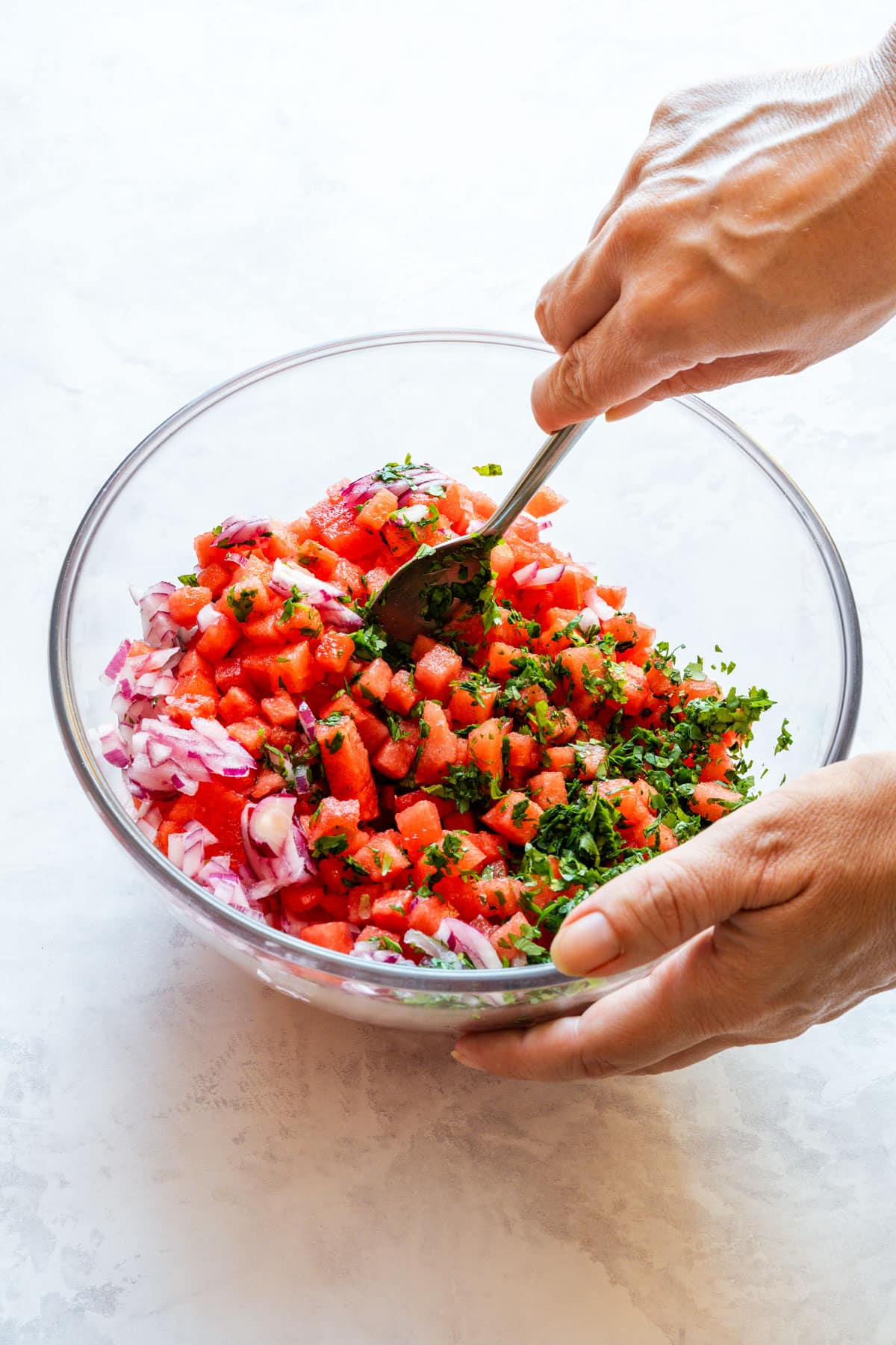 Watermelon, red onion and fresh herbs being tossed together in a glass bowl.