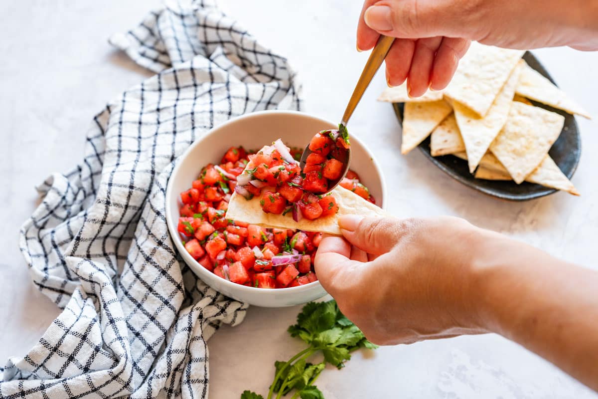 Spoonful of watermelon salsa being added to a tortilla chip, with more chips nearby.