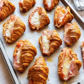 Mini ham and cheese croissants with melted Gruyère on a parchment-lined baking sheet.
