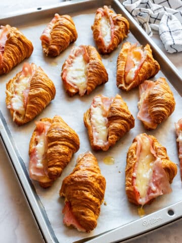 Mini ham and cheese croissants with melted Gruyère on a parchment-lined baking sheet.