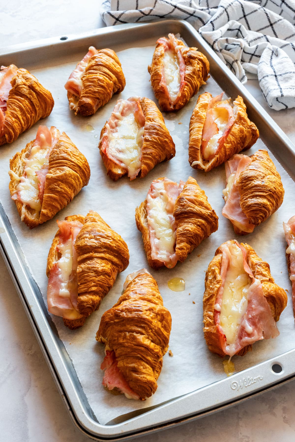 Mini ham and cheese croissants with melted Gruyère on a parchment-lined baking sheet.