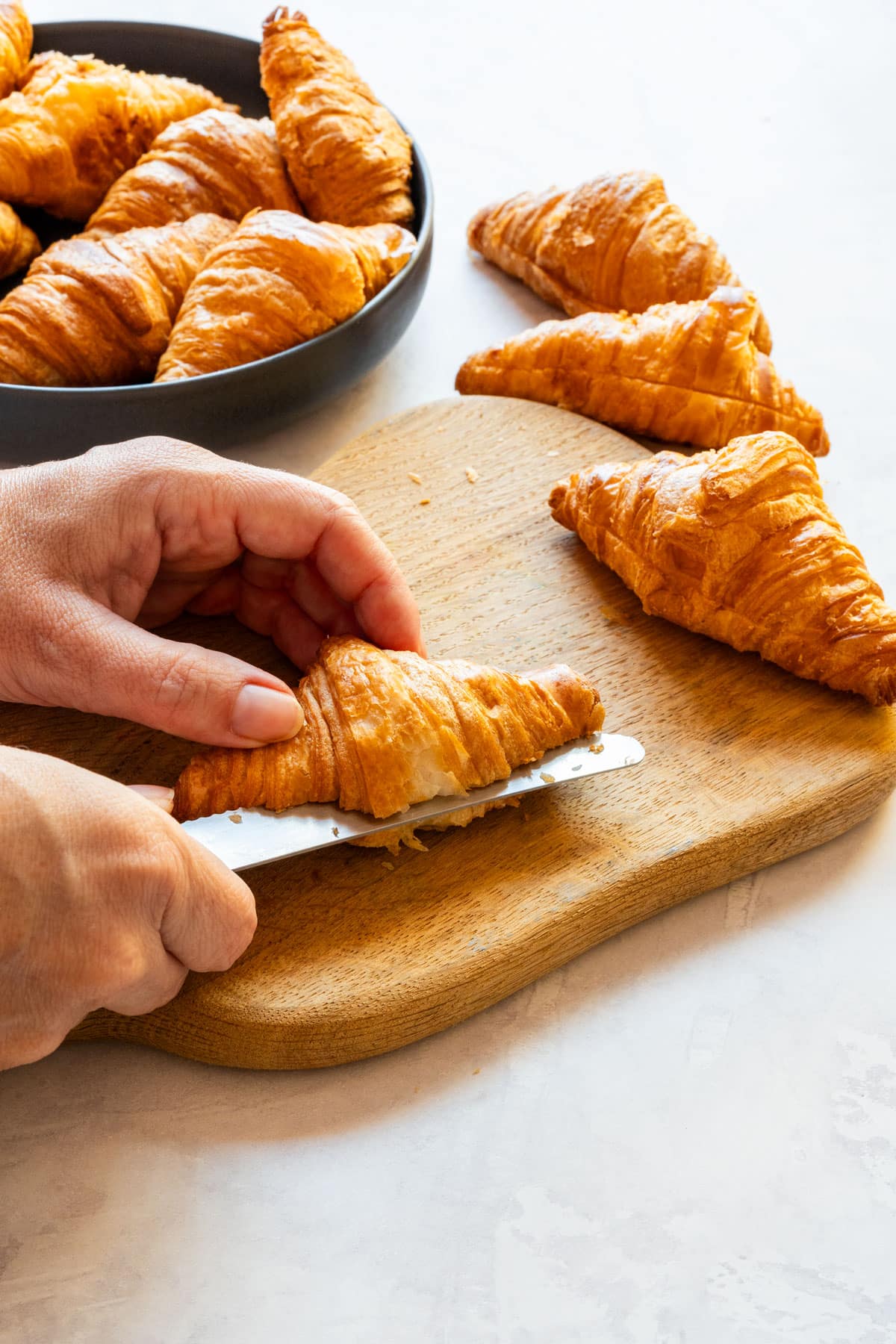 Cutting a mini croissant lengthwise with a serrated knife on a wooden board.