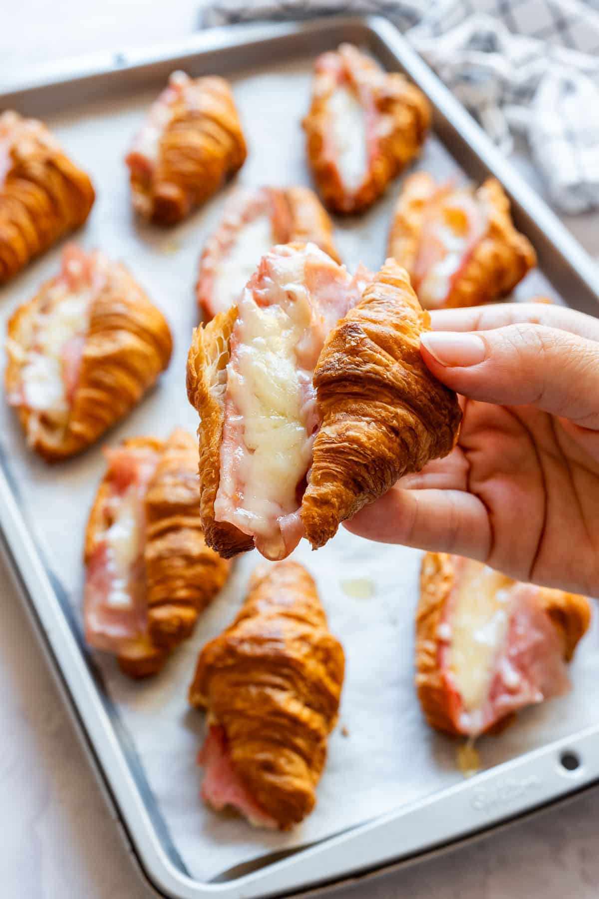 Hand holding a mini ham and cheese croissant above the tray, showing the melted filling inside.