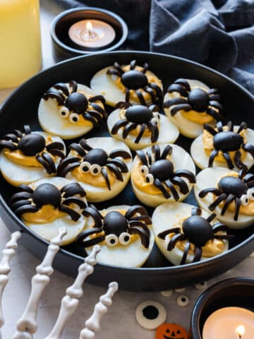 Spider deviled eggs made with black olives on a black plate, some with candy eyes, next to candles and a skeleton hand.