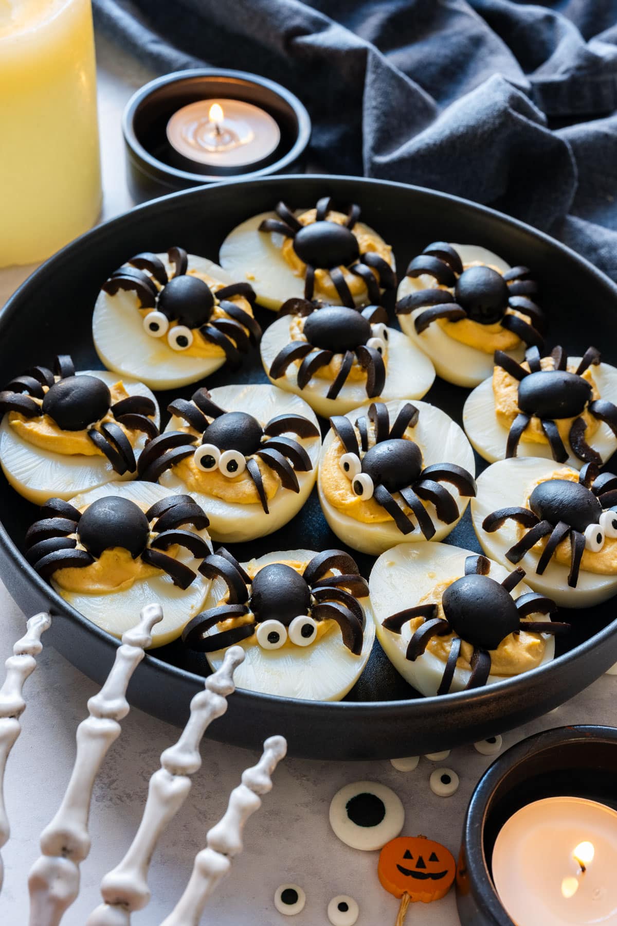 Spider deviled eggs made with black olives on a black plate, some with candy eyes, next to candles and a skeleton hand.