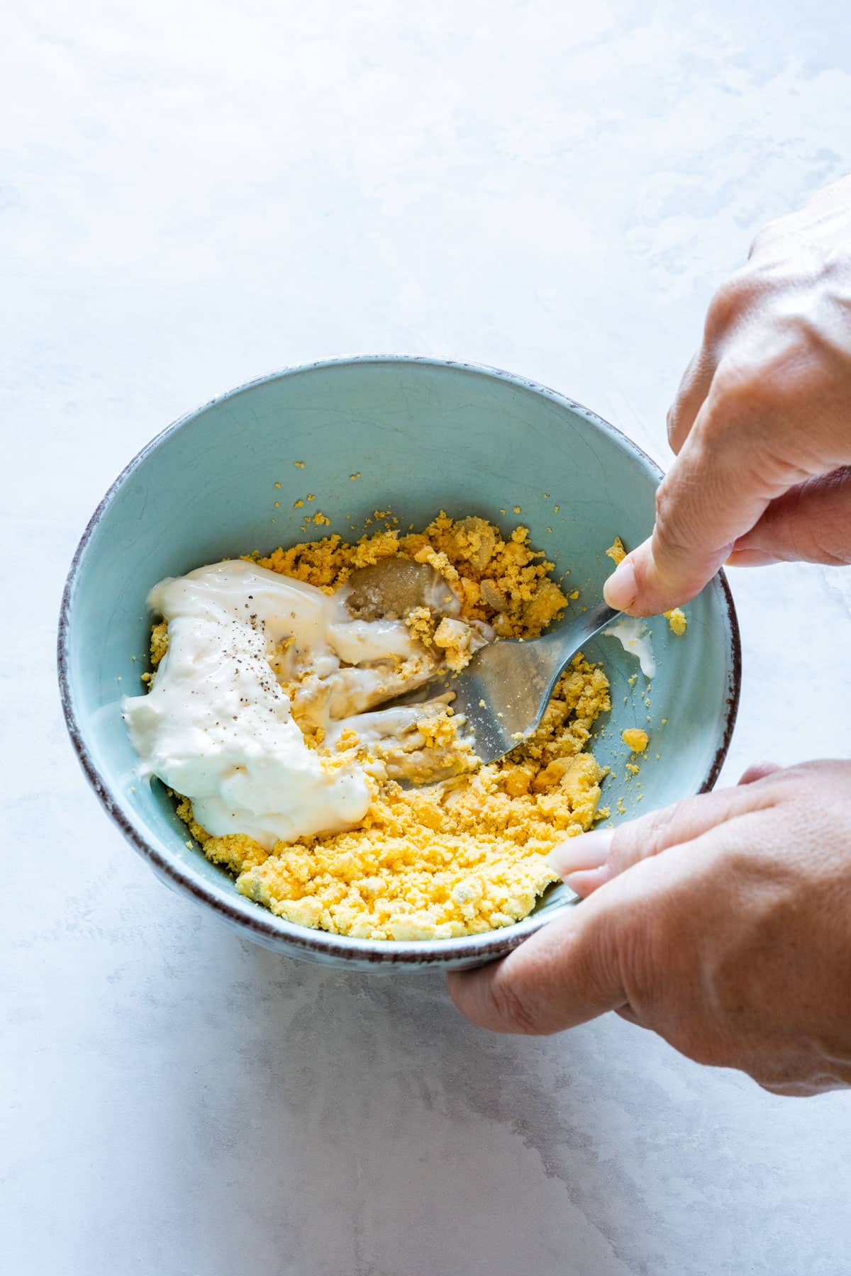 Mixing mashed egg yolks with mayonnaise, Dijon mustard and black pepper in a light blue bowl.