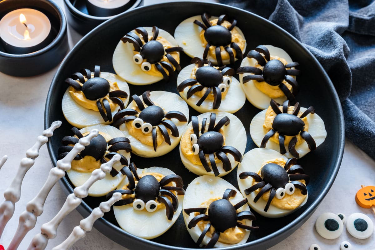 A plate of deviled eggs topped with black olive spiders, some with small candy eyes.
